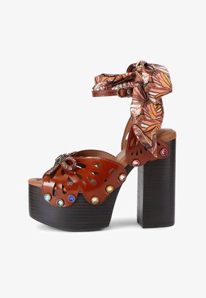 Brązowy skórzany sandał na platformie z wysokim, masywnym obcasem, wyciętym wzorem motyla, kolorowymi kamieniami szlachetnymi oraz ozdobnym paskiem wiązanym wokół kostki.