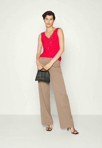 Mulher de top vermelho sem mangas com detalhes em renda, calças largas bege, bolsa preta e saltos altos pretos, em pé contra um fundo branco.