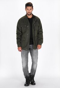 Giacca verde oliva con quattro tasche frontali, bottoni a pressione e una finitura texture, indossata sopra una maglietta nera e jeans grigi sbiaditi. Stivali neri.