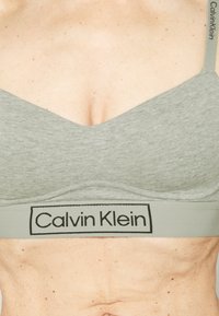 Pelēks kokvilnas bralets ar dziļu V-veida kakla izmēru un platu elastīgu jostu, uz kuras redzams melns Calvin Klein logotips. Gluda tekstūra un minimālisks stils.