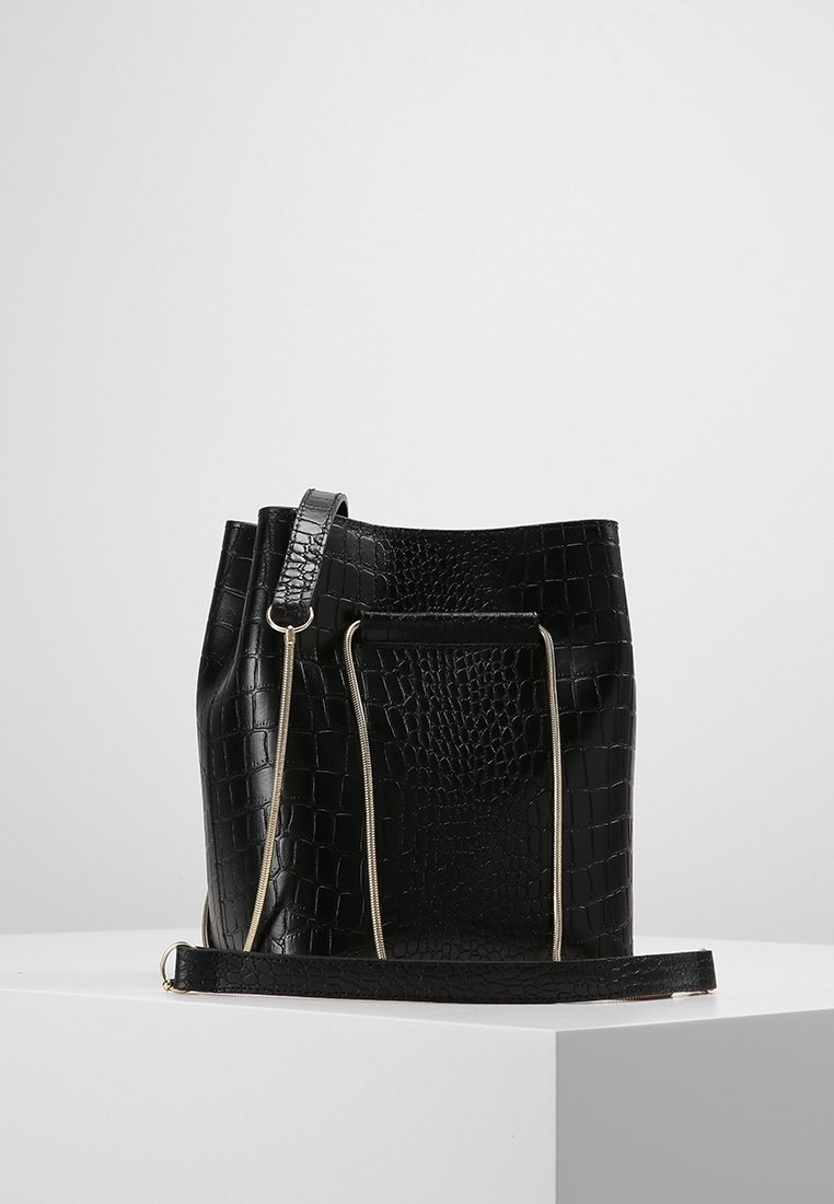 Sac seau en cuir noir embossé crocodile avec détails de fermeture éclair dorés et bandoulière détachable sur une surface blanche.
