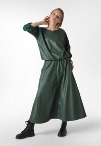 Top oversize de cuero verde combinado con una falda larga a juego, que presenta una cintura elástica y bolsillos laterales. Botas de combate negras con cordones.