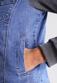 Denim vest with a light blue finish, featuring a hand in a front pocket. Gray fabric at the collar, creating a layered look.