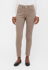 Beige skinny jeans med frontlommer og glidelås. Stoffet har en glatt tekstur, og designet har minimale sømdetaljer.