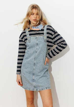 Femme blonde portant une chemise rayée à manches longues sous une robe salopette en jean bleu clair, debout avec une main sur la hanche.