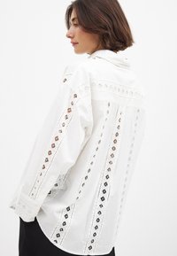 Camicia bianca con intricate decorazioni ricamate sulle spalle e sulle maniche, con bordi ondulati e polsini a bottoni.