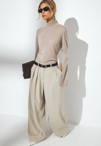 Beige turtleneck-toppi pitkillä hihoilla, yhdistettynä kermaisiin leveälahkeisiin housuihin. Musta vyö ja kudottu käsilaukku, viimeistelty terävillä koroilla.