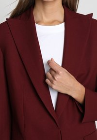 Kobieta w burgundowej marynarce na białej koszuli, trzymająca klapę marynarki prawą ręką w pobliżu klatki piersiowej.