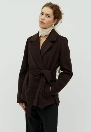 Femme portant un manteau marron foncé ceinturé, un pantalon noir, un foulard en dentelle blanc au cou et des boucles d'oreilles argentées en forme d’anneau sur un fond clair uni.