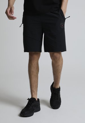 Mann iført svarte knelange shorts med en logo på høyre lår og svarte sportssko, stående mot en ensfarget bakgrunn.
