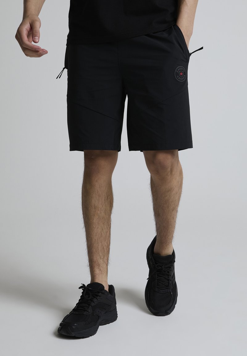 Homme portant un short noir mi-cuisses avec un logo sur la cuisse droite et des chaussures de sport noires, debout devant un fond uni.