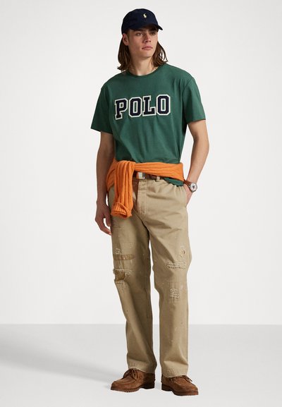 Groene katoenen T-shirt met 'POLO'-print, beige versleten broek, oranje trui om de taille geknoopt, marineblauwe pet en bruine schoenen.