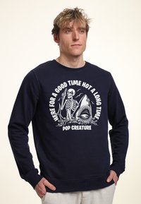 Sudadera navy de algodón, con un gráfico de un esqueleto y un tiburón; el texto dice "AQUÍ PARA PASAR UN BUEN RATO, NO UN RATO LARGO."