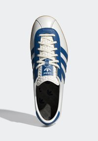 Vita Adidas-sneaker med blå ränder och tungslabel, avvit snörning och mönstrad innersula, visad ovanifrån på en ljus bakgrund.