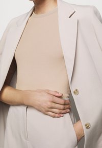 Gros plan d'une personne portant un haut côtelé beige, une veste et un pantalon beige clair, une main dans la poche et l'autre reposant sur la taille.