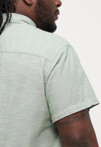 Cămașă cu mânecă scurtă, de culoare verde deschis, cu un croi lejer, dintr-un material texturat și cu guler; detalii de cusătură vizibile și un tatuaj pe braț.