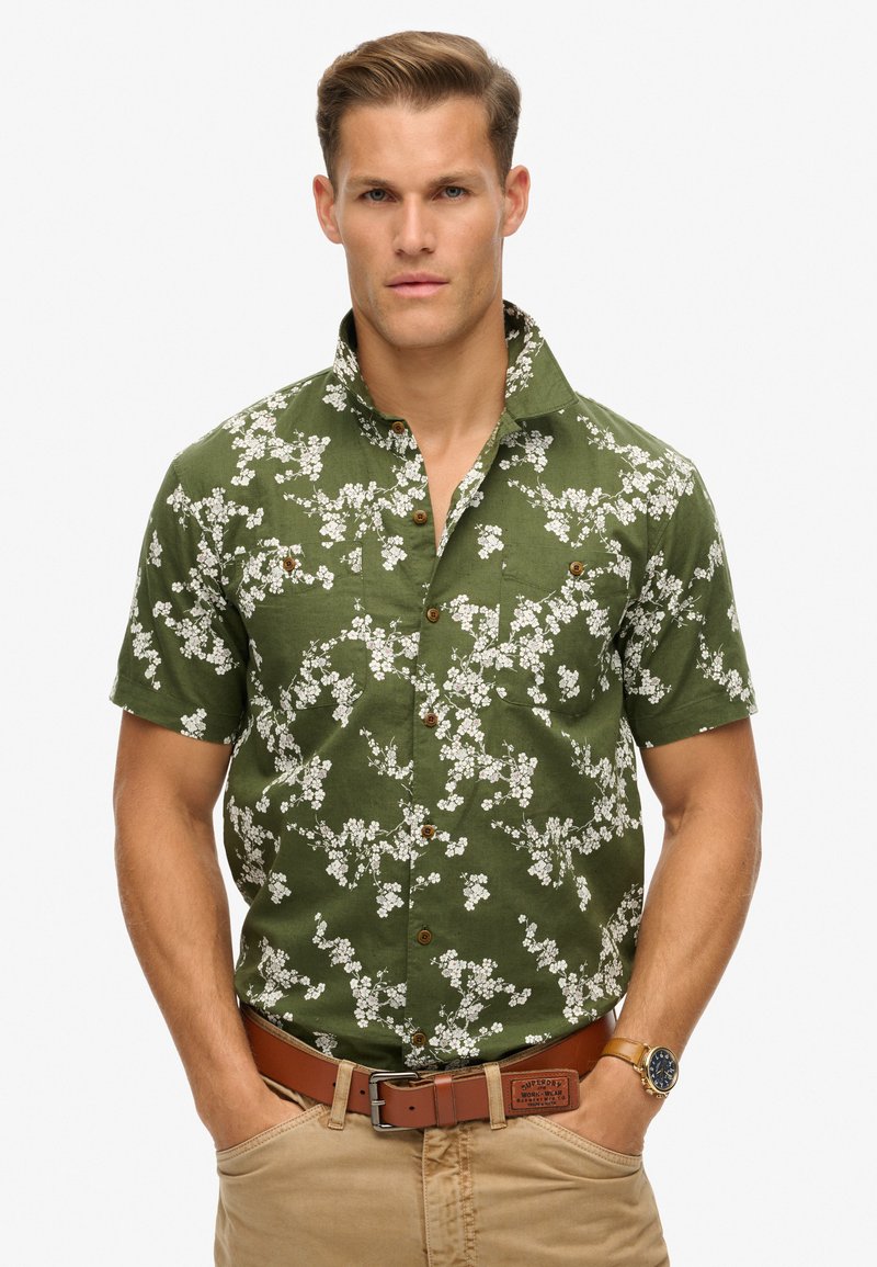 Camisa de manga corta de color verde oliva con un patrón floral, diseño de botones y dos bolsillos en el pecho. Presenta botones marrones y un cuello estructurado.
