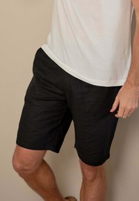 Mand iført sorte shorts og en hvid t-shirt, stående mod en ensfarvet beige baggrund, som viser en del af hans overkrop og ben.