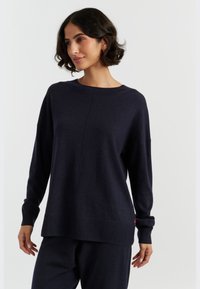 Marineblauer Strickpullover mit rundem Ausschnitt, langen Ärmeln, herunterhängenden Schultern und geripptem Saum; zeichnet sich durch einen dezenten roten Akzent an der Seite aus.