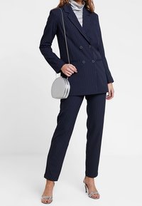 Marineblauer gestreifter, doppelt geknöpfter Blazer und passendes, tailliertes Hosen, kombiniert mit einer silbernen, runden Handtasche und metallicfarbigen High Heels.
