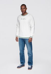 Vit sweatshirt med grön ribbad krage och muddar, med en logotyp i skriftsnitt. Kombinerad med blå denimjeans och marinblå sneakers med vita skosnören.
