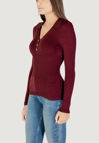 Pullover bordeaux a maniche lunghe con colletto, scollatura abbottonata, dettaglio a coste e vita aderente, abbinato a jeans azzurri chiari.