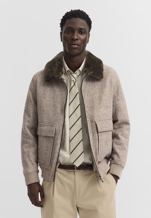 Junger Mann trägt beige Jacke mit Fellkragen, gestreiftes Hemd, passender gestreifter Krawatte, helle Hose und braunen geflochtenen Gürtel.