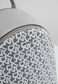 Grå ryggsäck med mönstrad framsida som har sammanlänkade geometriska former i vitt och svart, texturerat material och silverdetaljer på dragkedjan.