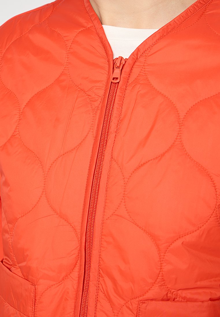 Pikowana pomarańczowa kurtka z zamkiem błyskawicznym z przodu, charakteryzująca się gładką teksturą oraz okrągłymi wzorami szwów, a także bocznymi kieszeniami.