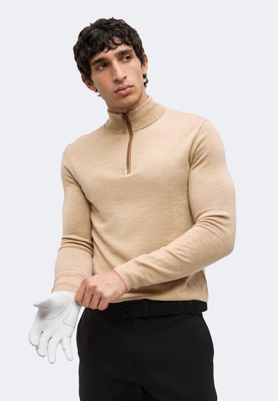 Jeune homme portant un pull beige zippé et un pantalon noir met un gant blanc, regardant sur le côté sur un fond clair uni.