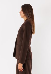 Blazer marrone su misura con maniche lunghe, una silhouette elegante e una tasca singola. Texture liscia e design moderno, accompagnato da pantaloni abbinati.