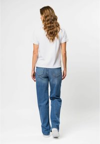 Witte katoenen t-shirt met korte mouwen, gecombineerd met lichtblauwe, iets versleten denim jeans en witte sneakers, gezien van achteren.