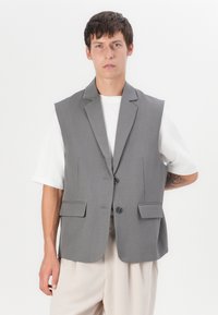 Šedý bezrukávový blazer s klopy, dvěma předními kapsami a dvěma knoflíky. Nositý přes bílou košili, která má hladkou texturu.