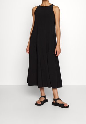 Kobieta ubrana w czarną, bezrękawnikową, luźną sukienkę midi, w połączeniu z czarnymi sandałami na platformie, stojąca na tle jednolitego białego tła.