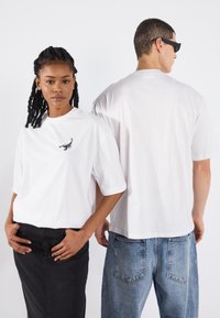 Vita kortärmade t-shirts med en svart skorpionbroderi på framsidan. Modellen har på sig en svart kjol; den andra har blå jeans.