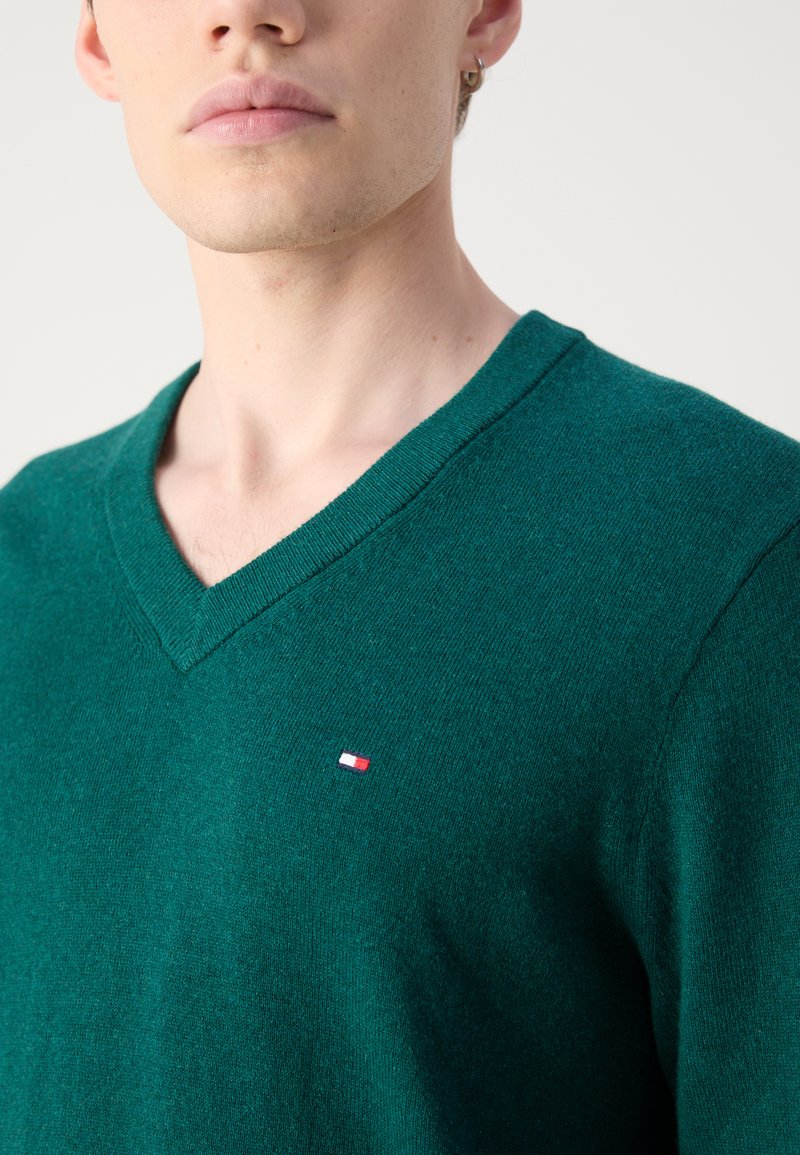 Zielony sweter w serek wykonany z miękkiego dzianiny, z małym czerwono-biało-niebieskim logo po lewej stronie klatki piersiowej. Zbliżenie na dekolt.
