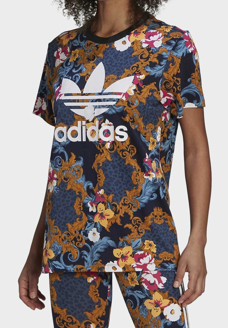Floral-patterned navy t-shirt with gold accents, featuring a white Adidas logo. Short sleeves and a relaxed fit. Made from a lightweight material.