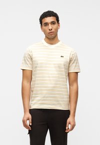 Άνδρας που φοράει μπλούζα κοντομάνικη με μπεζ και λευκές ρίγες και μικρό λογότυπο στο στήθος, συνδυασμένη με μαύρο παντελόνι, στέκεται μπροστά σε λευκό φόντο.