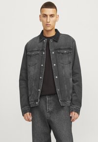 Giubbotto in denim nero con una finitura leggermente sbiadita, chiusura con bottoni, due tasche sul petto e un colletto. Indossato sopra una maglietta nera.