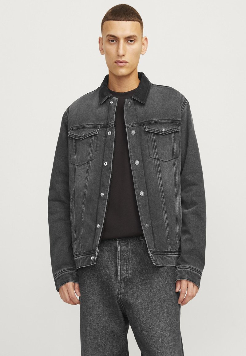 Giubbotto in denim nero con una finitura leggermente sbiadita, chiusura con bottoni, due tasche sul petto e un colletto. Indossato sopra una maglietta nera.