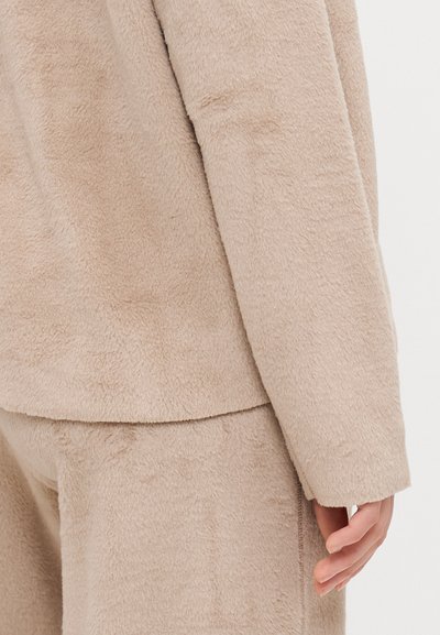 Beige fleece garment featuring soft texture, loose fit, and minimal seams. The sleeve extends comfortably over the hand, showcasing its relaxed design.
