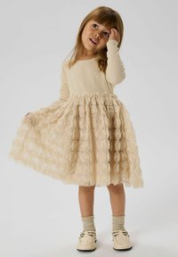 Beige langärmliges Oberteil mit einem rüschierten Bund, kombiniert mit einem creamfarbenen Rock mit Rosenmuster in Schichten. Weiße Schuhe mit Schnallendetail und strukturierten Socken.