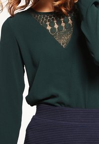 Blouse vert foncé à manches longues, avec un détail en dentelle au niveau du col, accompagnée d'une jupe bleu marine texturée et côtelée en dessous.
