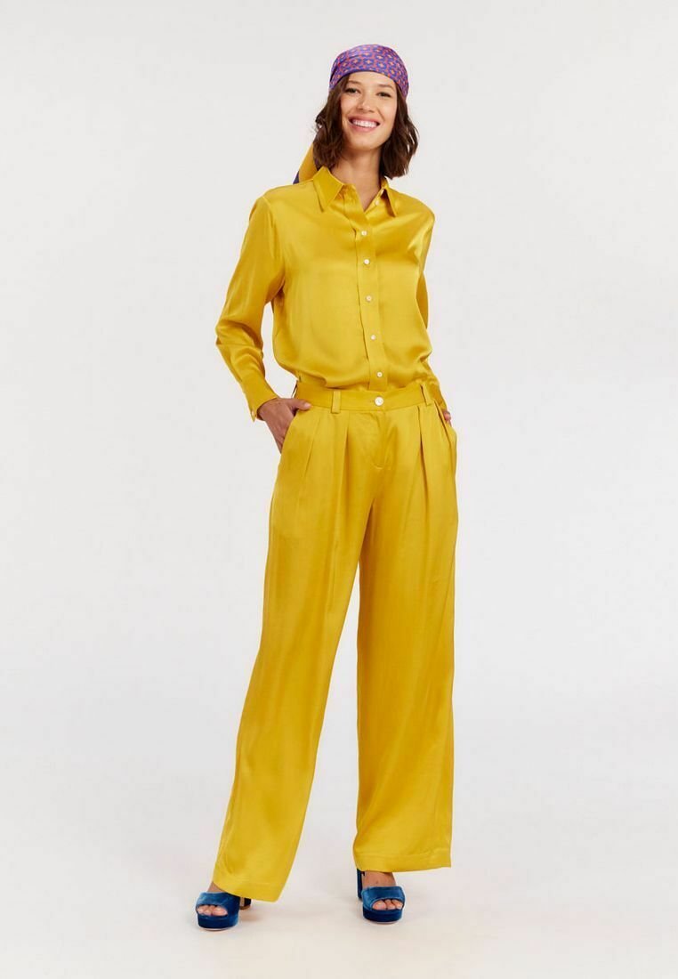 Blouse en soie jaune avec des boutons, associée à un pantalon plissé taille haute assorti, et des sandales à plateforme ouvertes bleues. Foulard à motifs.