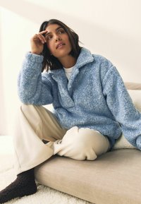 Lyseblå fleecepullover med krave, båret over en hvid skjorte og lys beige fløjlsbukser. Mørkebrune fuzzy sokker på en beige sofa.