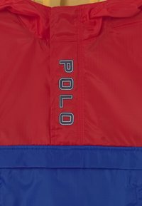 Červený a modrý lehký větrovka s kapucí, na červeném panelu je nápis "POLO" bílými písmeny. Hladký povrch, syntetický materiál.