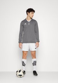 Grijze sporttop met lange mouwen en een halve rits, witte shorts, zwarte voetbalschoenen en zwarte gestreepte sokken, staande naast een voetbal.