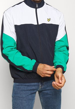 Mand, der justerer ærmet på en blå, hvid og turkis lynlåstrøje med en lille gul fuglelogo på brystet, iført blå jeans.