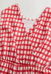 Rødt og hvidt ternet stof med V-hals og elastisk taljebånd, der viser tekstureret, let materiale og nærbillede af syningsdetaljer.