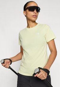 Ανοιχτό κίτρινο κοντομάνικο μπλουζάκι με στρογγυλή λαιμόκοψη και μικρό λευκό λογότυπο. Μαύρα ρυθμιζόμενα γυαλιά ηλίου. Ο χρήστης κρατά μπαστούνια πεζοπορίας.